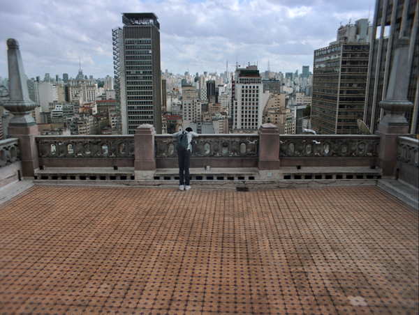 Cena de Cidade Pássaro com o protagonista sozinho em um grande terraço com São Paulo ao fundo.