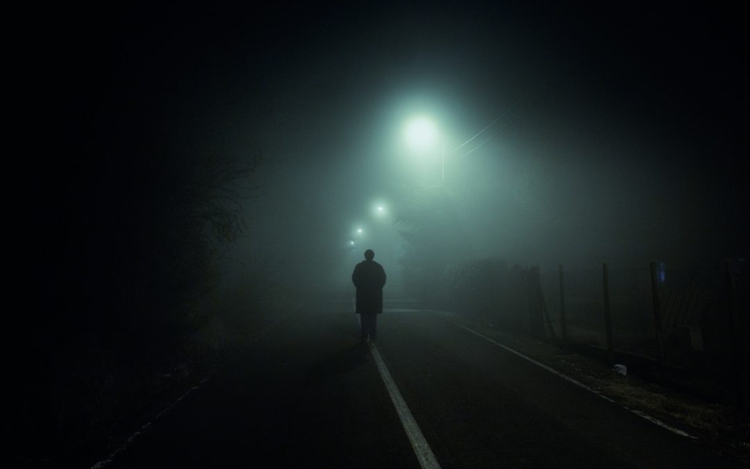 Foto noturna, escura e enevoada com uma pessoa indo embora por uma estrada e sumindo na névoa.